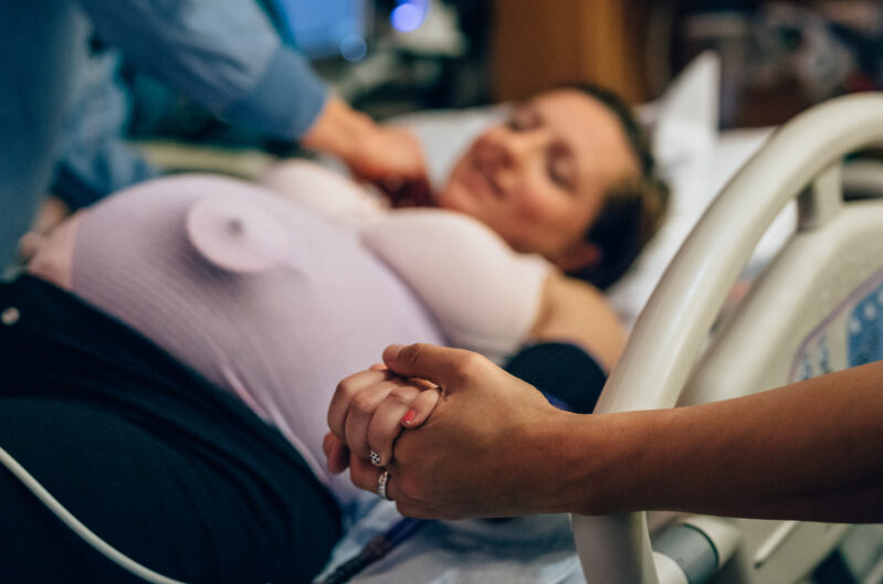 A fetal monitor is visible on a pregnant mother's belly as someone holds her hand